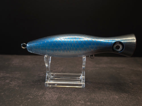 A metallic black and blue fishing lure with a red mouth, displayed on a clear acrylic stand against a dark background.