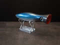 A metallic black and blue fishing lure with a red mouth, displayed on a clear acrylic stand against a dark background.