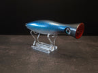 A metallic black and blue fishing lure with a red mouth, displayed on a clear acrylic stand against a dark background.
