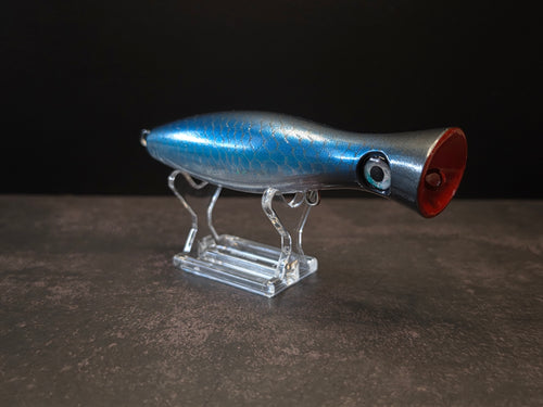 A metallic black and blue fishing lure with a red mouth, displayed on a clear acrylic stand against a dark background.