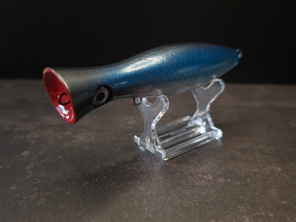 A metallic black and blue fishing lure with a red mouth, displayed on a clear acrylic stand against a dark background.
