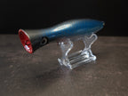 A metallic black and blue fishing lure with a red mouth, displayed on a clear acrylic stand against a dark background.