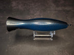 A metallic black and blue fishing lure with a red mouth, displayed on a clear acrylic stand against a dark background.