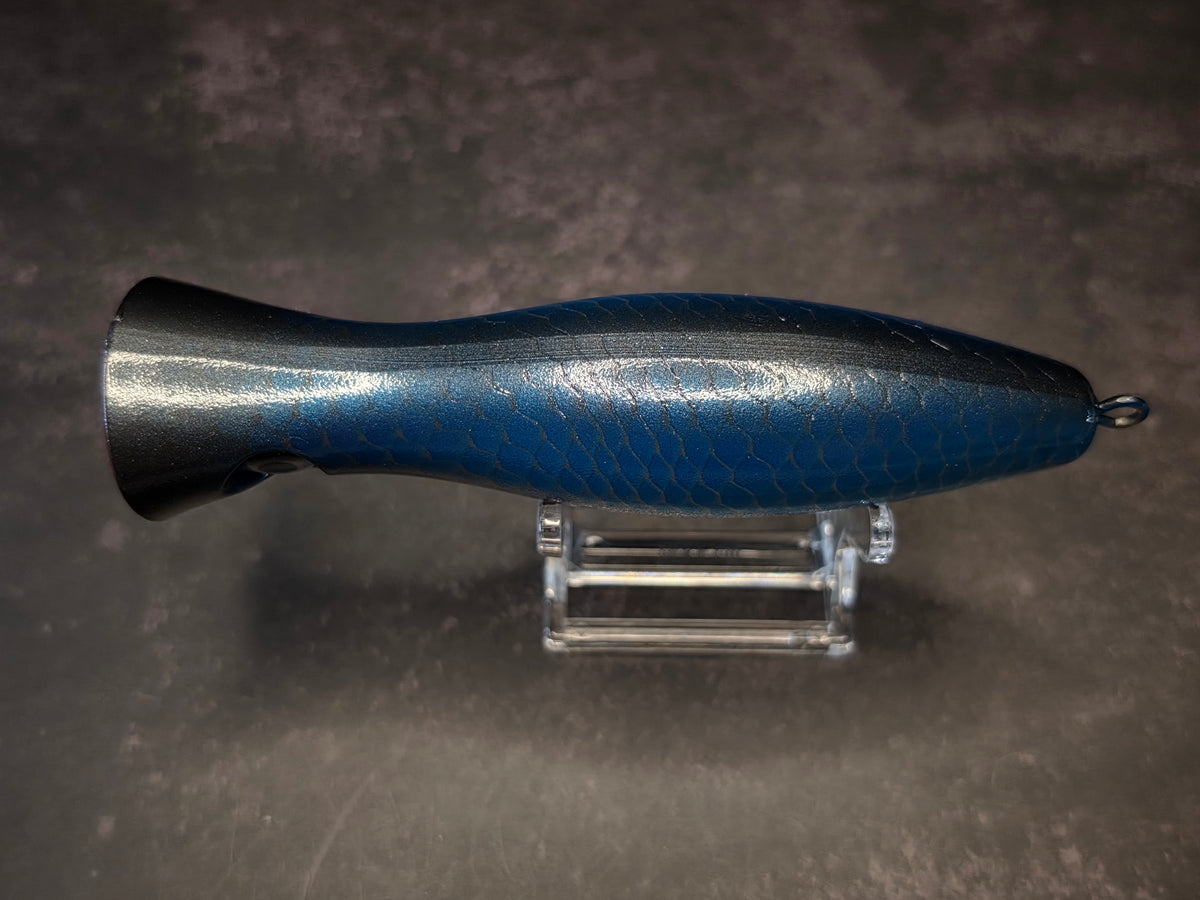 A metallic black and blue fishing lure with a red mouth, displayed on a clear acrylic stand against a dark background.