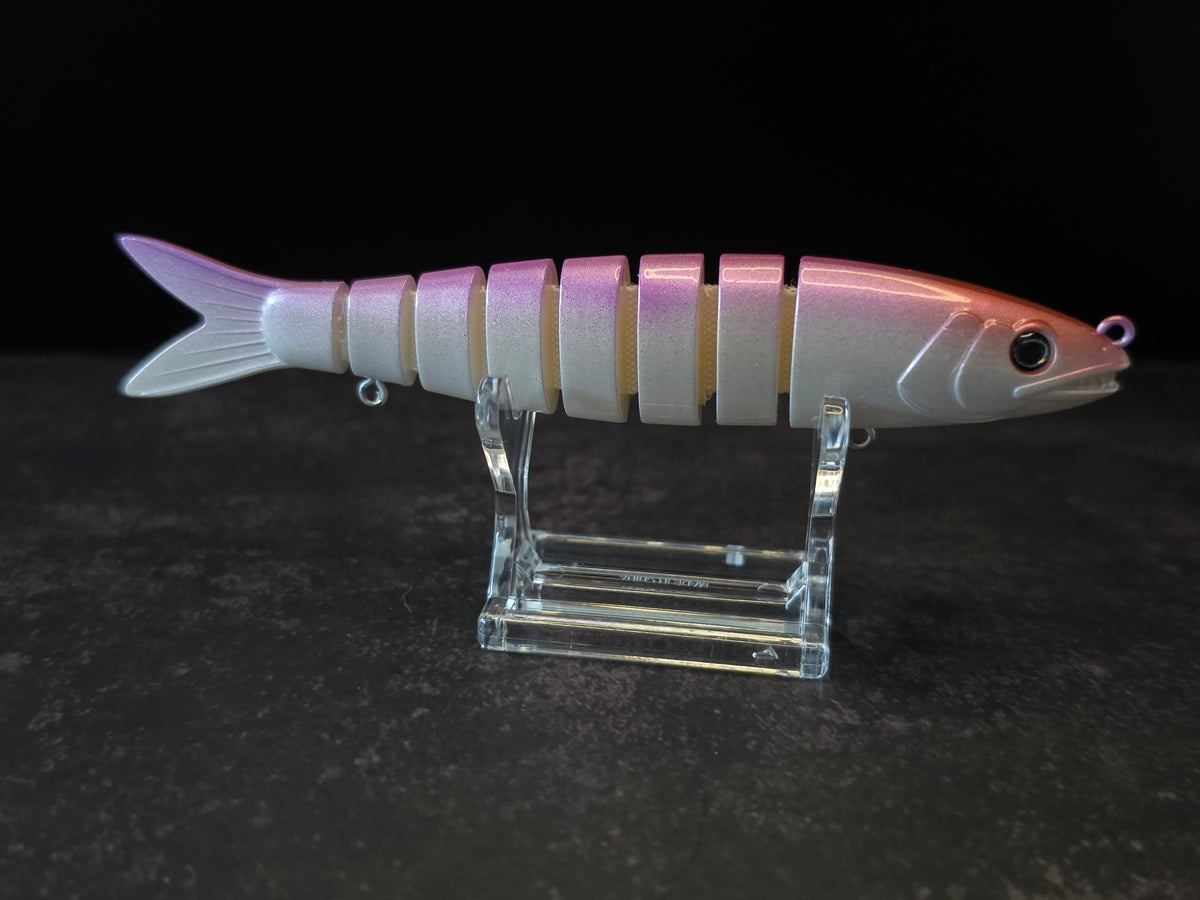 A pink and white gradient hard fishing lure shaped like a small fish, displayed on a clear acrylic stand against a dark background.