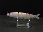 A pink and white gradient hard fishing lure shaped like a small fish, displayed on a clear acrylic stand against a dark background.