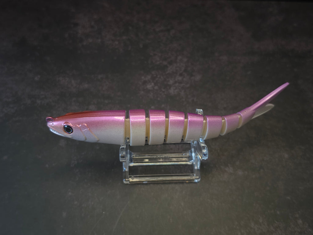 A pink and white gradient hard fishing lure shaped like a small fish, displayed on a clear acrylic stand against a dark background.