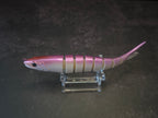 A pink and white gradient hard fishing lure shaped like a small fish, displayed on a clear acrylic stand against a dark background.