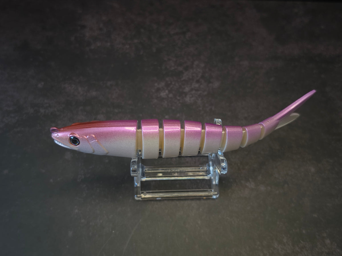 A pink and white gradient hard fishing lure shaped like a small fish, displayed on a clear acrylic stand against a dark background.