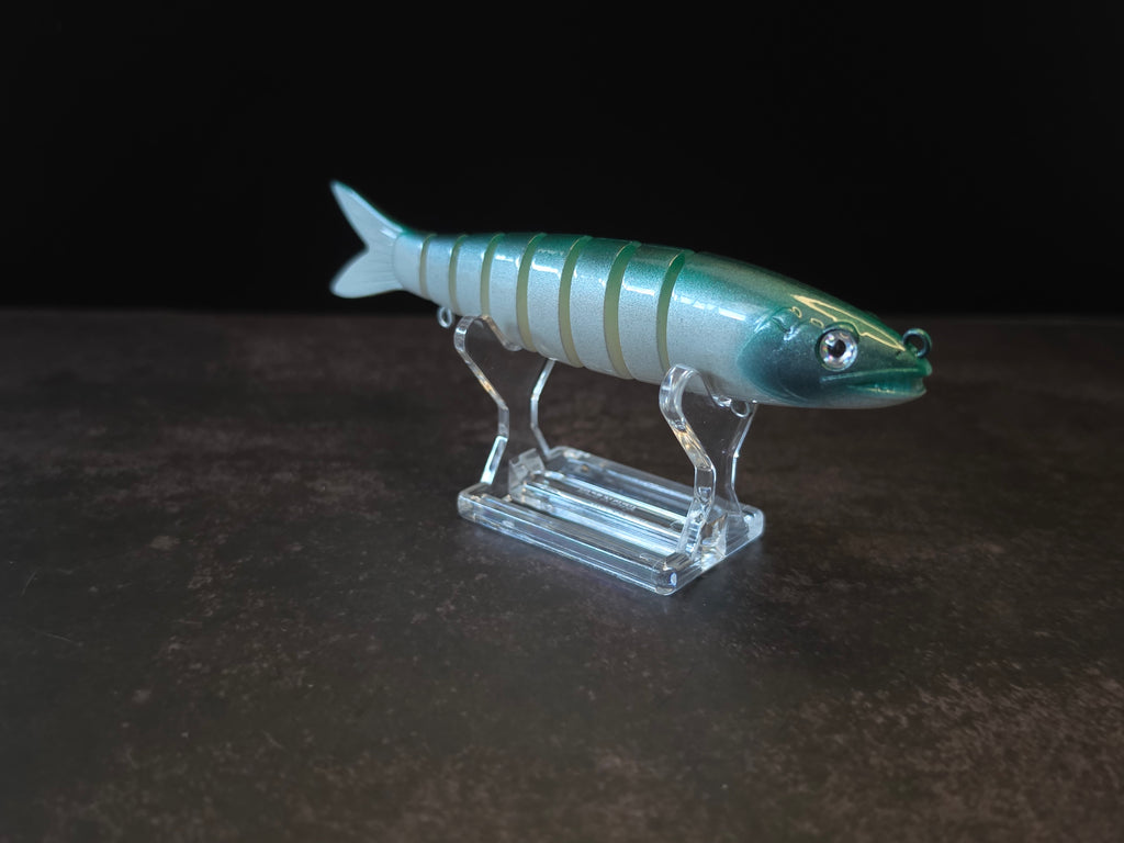 A hard plastic fishing lure shaped like a fish, featuring green to white gradient coloring with a green head, displayed on a clear acrylic stand against a dark background.