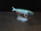 A hard plastic fishing lure shaped like a fish, featuring green to white gradient coloring with a green head, displayed on a clear acrylic stand against a dark background.