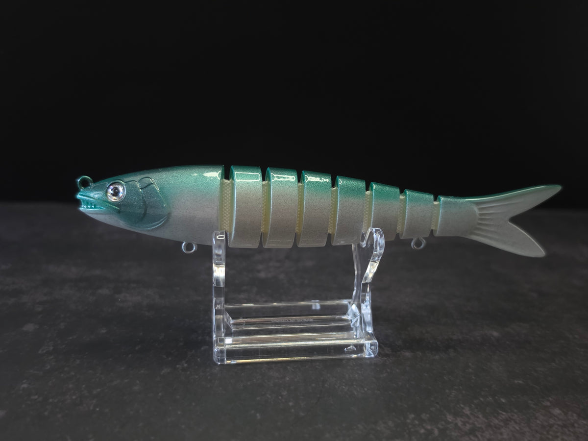 A hard plastic fishing lure shaped like a fish, featuring green to white gradient coloring with a green head, displayed on a clear acrylic stand against a dark background.