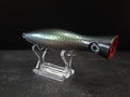 A metallic black and green fishing lure with a red mouth, displayed on a clear acrylic stand against a dark background.