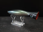 A metallic black and green fishing lure with a red mouth, displayed on a clear acrylic stand against a dark background.