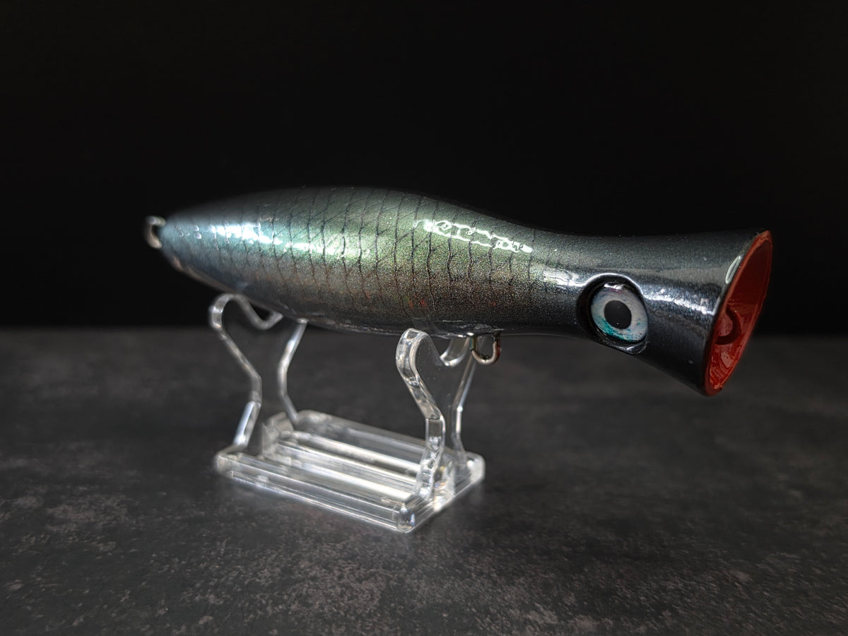 A metallic black and green fishing lure with a red mouth, displayed on a clear acrylic stand against a dark background.