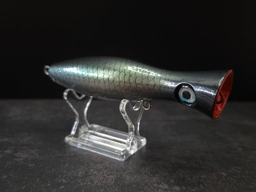 A metallic black and green fishing lure with a red mouth, displayed on a clear acrylic stand against a dark background.