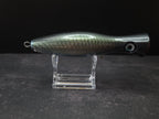 A metallic black and green fishing lure with a red mouth, displayed on a clear acrylic stand against a dark background.