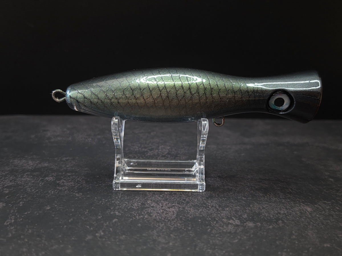 A metallic black and green fishing lure with a red mouth, displayed on a clear acrylic stand against a dark background.