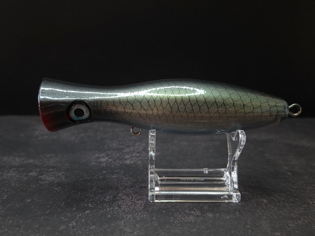 A metallic black and green fishing lure with a red mouth, displayed on a clear acrylic stand against a dark background.