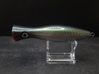 A metallic black and green fishing lure with a red mouth, displayed on a clear acrylic stand against a dark background.