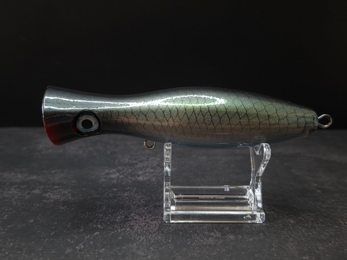 A metallic black and green fishing lure with a red mouth, displayed on a clear acrylic stand against a dark background.