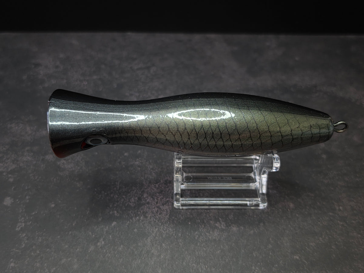 A metallic black and green fishing lure with a red mouth, displayed on a clear acrylic stand against a dark background.